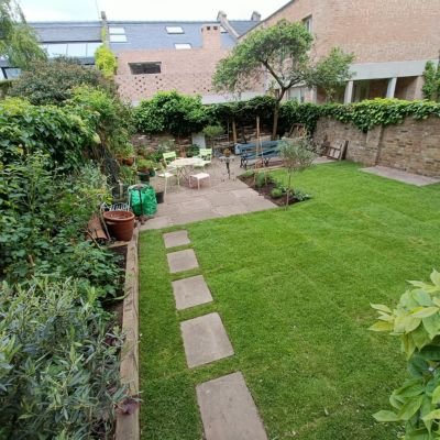 A lush, vibrant green lawn in a private residential courtyard featuring square stone stepping stones, garden beds, and patio furniture, demonstrating the results of professional lawn feeding.