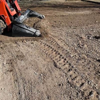 A close-up of an orange skid steer loader bucket grading and leveling dry, rocky topsoil, leaving visible tire tracks in the dirt as it prepares the ground for a new lawn.