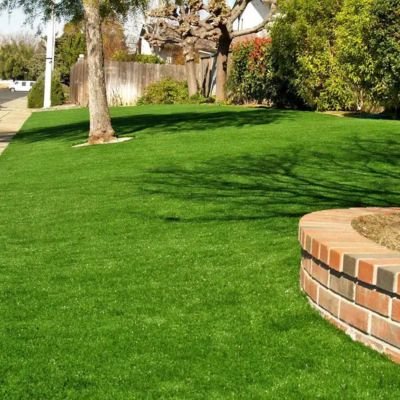 A wide-angle landscape shot of a newly installed, vibrant green sod lawn covering a front yard slope, framed by a concrete sidewalk, mature trees, and a low curved brick retaining wall.
